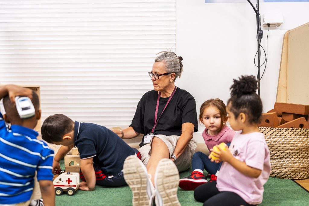 A teacher sat on the floor with four students, showing interest in their activities